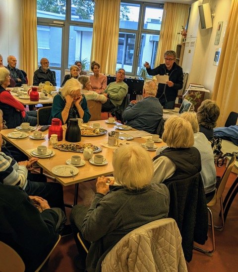 Mehrere Personen sitzen bei Kaffee und Kuchen und hören einem Vorleser zu