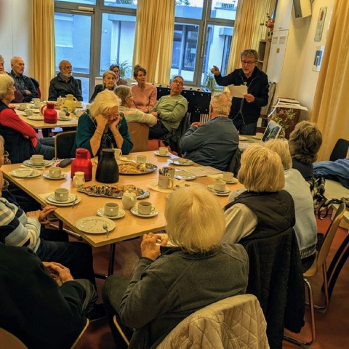Mehrere Personen sitzen bei Kaffee und Kuchen und hören einem Vorleser zu